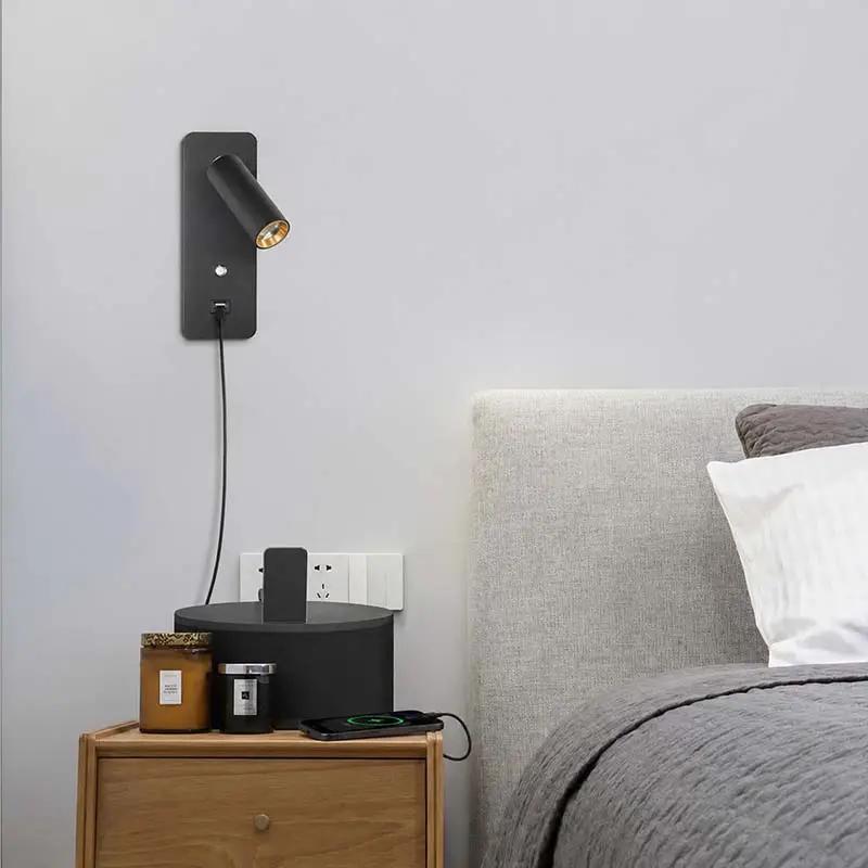 A modern black wall-mounted spotlight with a gold interior trim, shown turned on and plugged into a bedside table in a bedroom setting next to a bed.