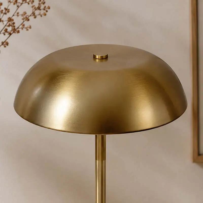 A modern mushroom-shaped table lamp with a brushed brass dome shade and a slim matching stem, shown against a warm beige background.